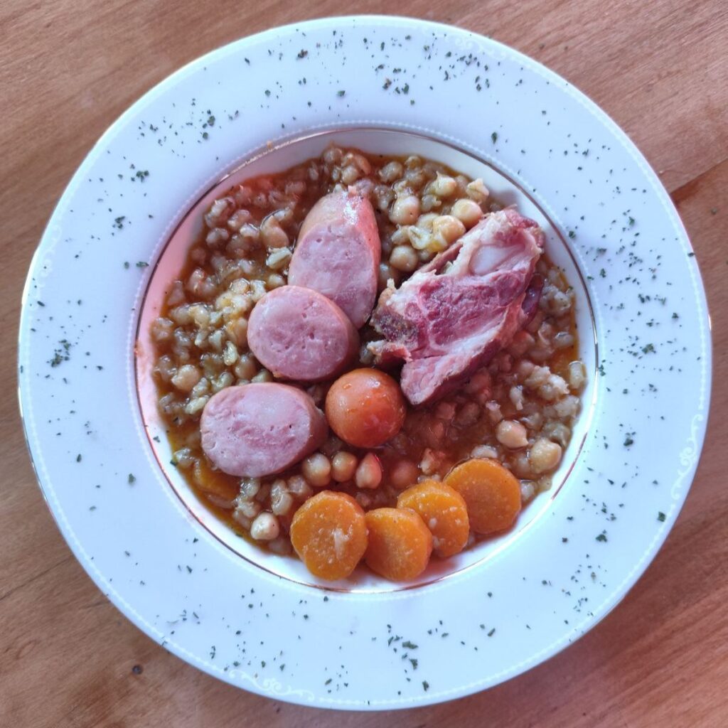 A plate of Barley Chickpea Meat Stew served with sliced smoked pork, sausage rounds, cherry tomato, and carrot garnish.