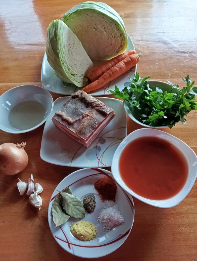 All ingredients for Savory Cabbage Pancetta Stew arranged whole on a wooden table, including cabbage, carrots, pancetta, onion, garlic, celery, parsley, oil, tomato passata, bay leaves, paprika, salt, Vegeta, and black pepper.