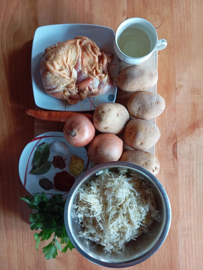 All ingredients for Cabbage Chicken Potato Medley arranged on a table, including sauerkraut, chicken drumsticks, potatoes, onions, carrot, oil, spices, and herbs.