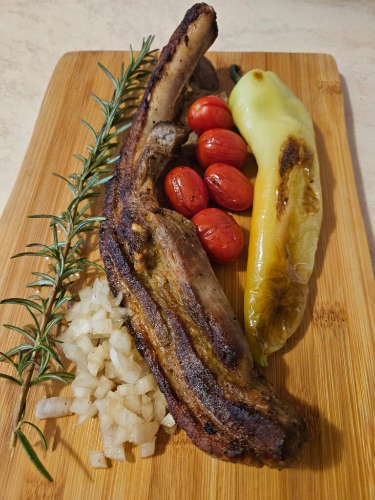 Served pork rib on a wooden board with grilled pepper, cherry tomatoes, chopped onion, and rosemary decoration.