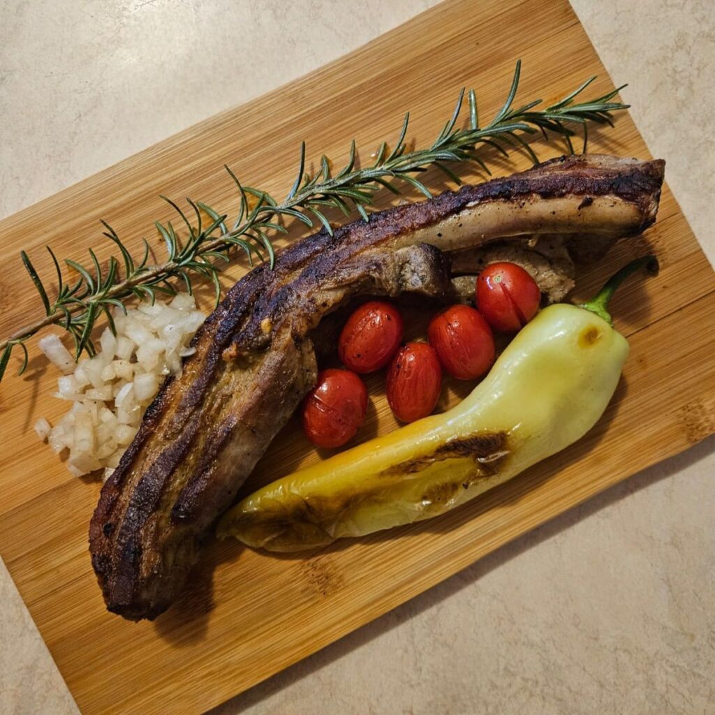 One wooden board with a grilled pork rib, a grilled pepper, several cherry tomatoes, chopped onion, and a rosemary sprig.