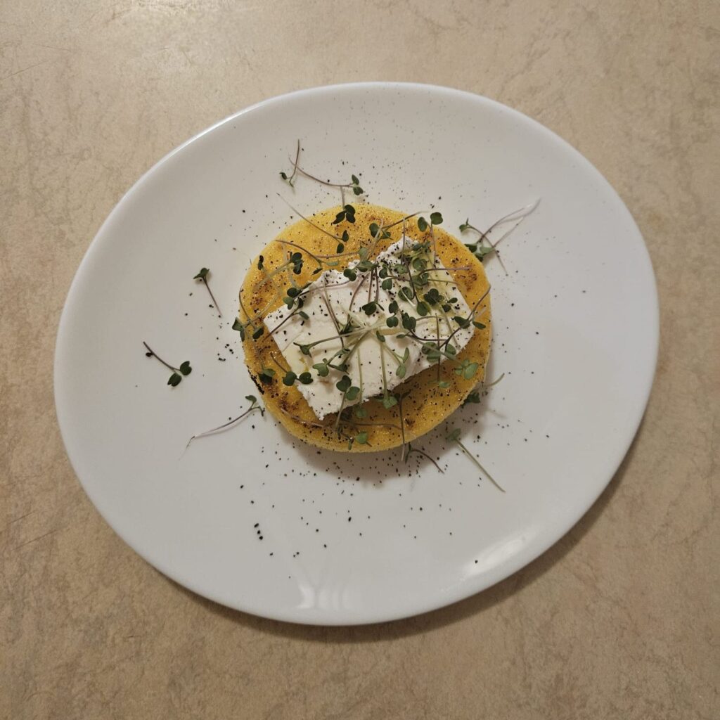 Served polenta round topped with goat cheese, drizzled with olive oil, seasoned with pepper, and garnished with arugula microgreens.