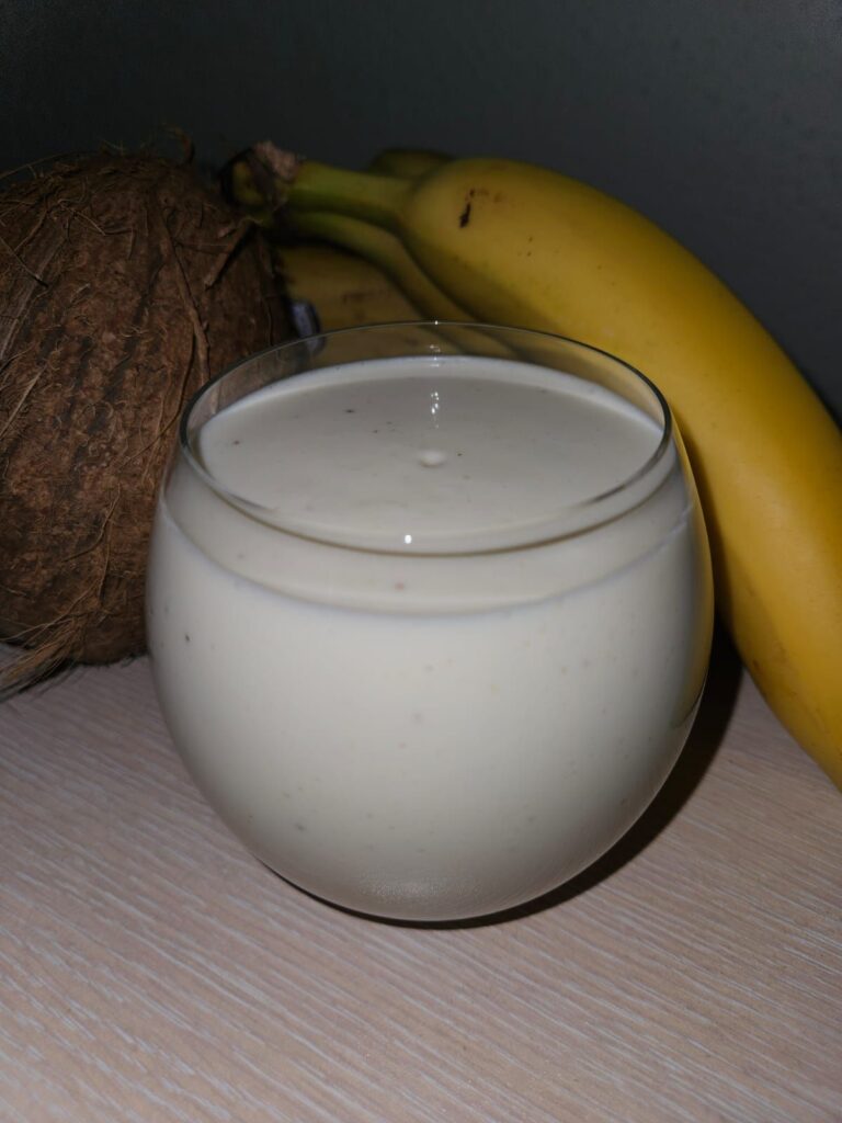 Refreshing Banana Hazelnuts Coconut Smoothie served in a glass with bananas and a whole coconut in the background.