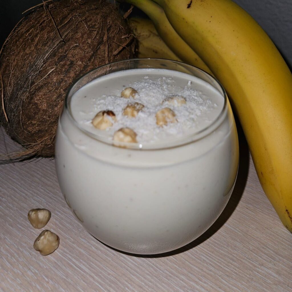 Banana Hazelnuts Coconut Smoothie served in a glass topped with hazelnuts and coconut flakes, with bananas and a coconut behind.