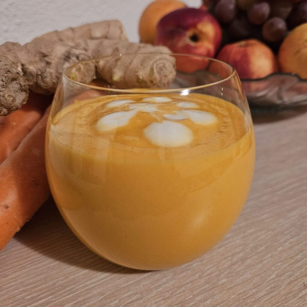 Glass of Apricot Carrot Ginger Smoothie with fresh carrots, ginger, and colorful fruits in the background.