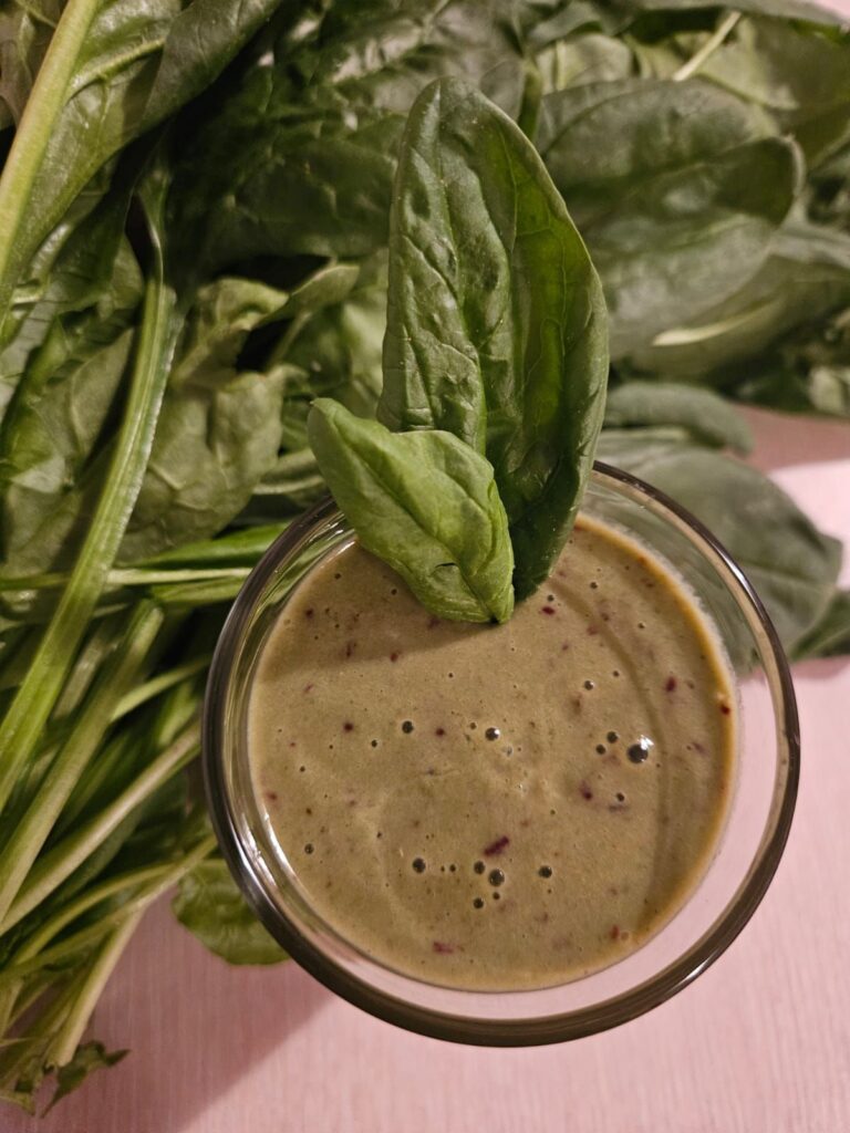 Overhead view of Black Currants Spinach Smoothie in a glass with fresh spinach leaves beside it.