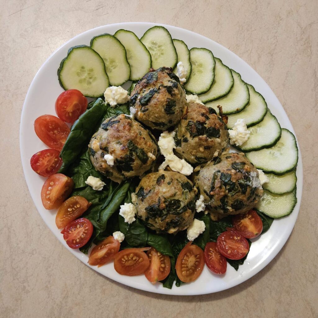 A plate of baked turkey meatballs with spinach and feta, served with fresh tomatoes, cucumber slices, and spinach.