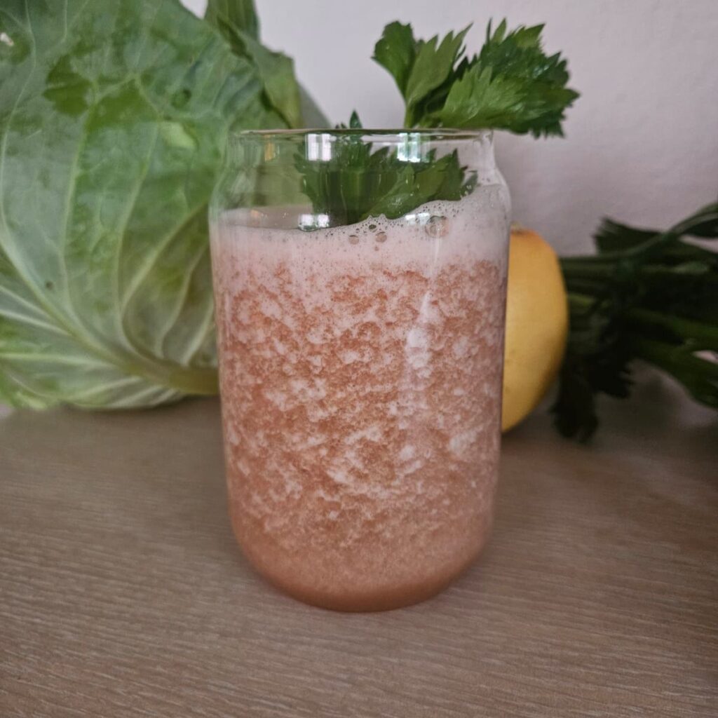 Glass of grapefruit celery cabbage juice decorated with celery leaves, with whole cabbage, grapefruit, and celery stalks in the background.