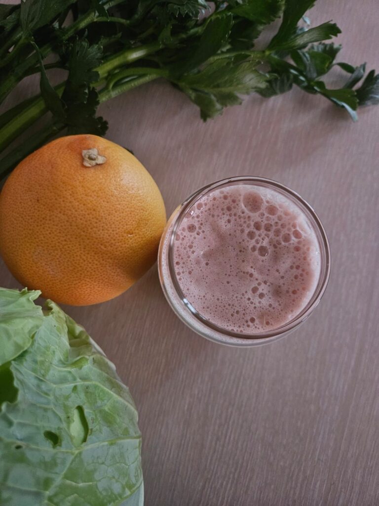 Top view of a glass filled with grapefruit celery cabbage juice, surrounded by whole cabbage, grapefruit, and fresh celery stalks with leaves.