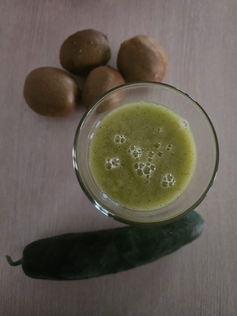 Top view of a glass filled with kiwi mango celery juice, with a few whole kiwi and one cucumber in the background.