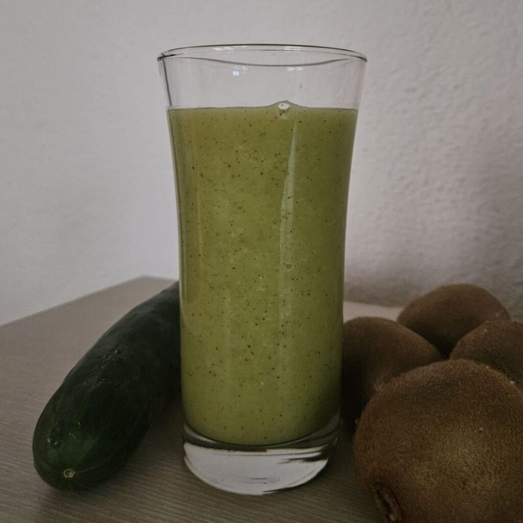 Glass of kiwi mango celery juice placed in front of a few whole kiwi and one cucumber.