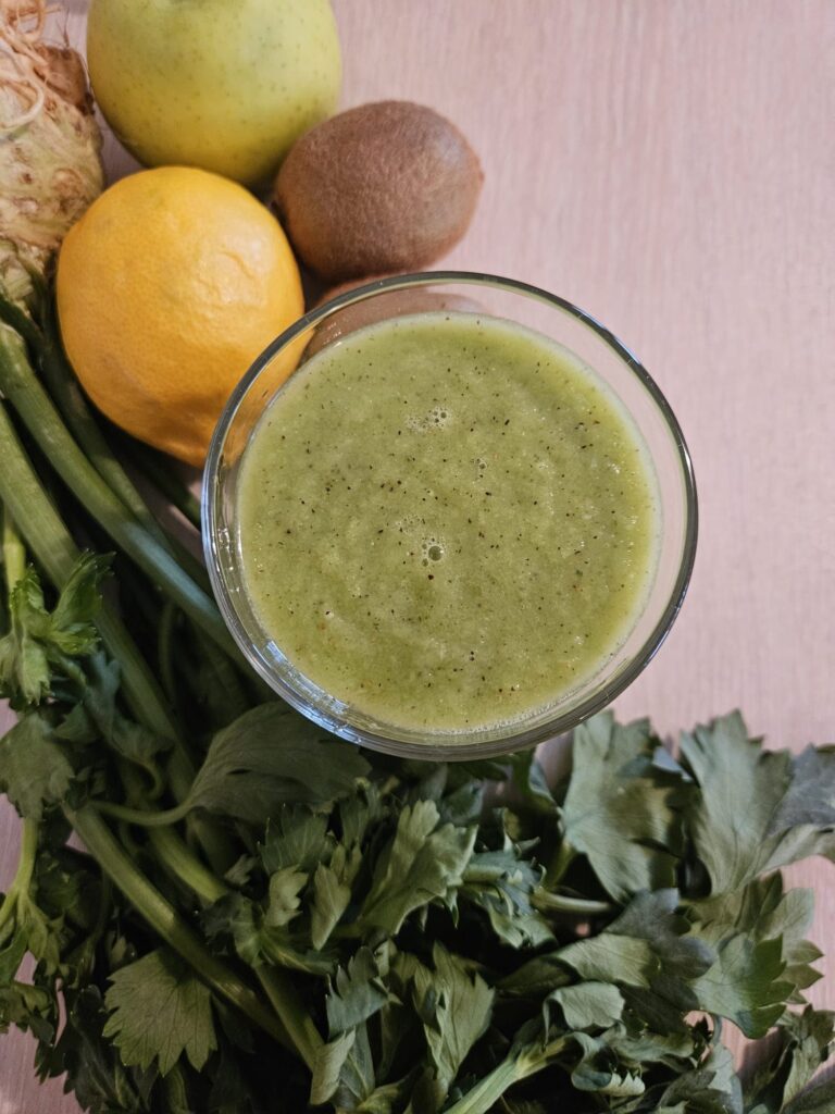 Top view of a fresh apple kiwi celery juice in a glass with celery, kiwi, green apple, and lemon in the background.