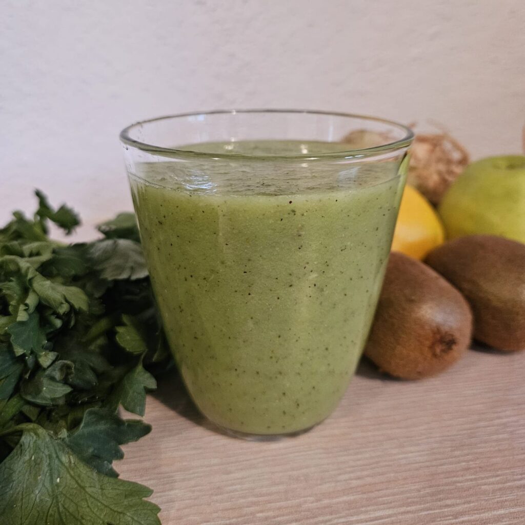 Side view of apple kiwi celery juice in a glass with green apple, kiwi, celery, and lemon in the background.