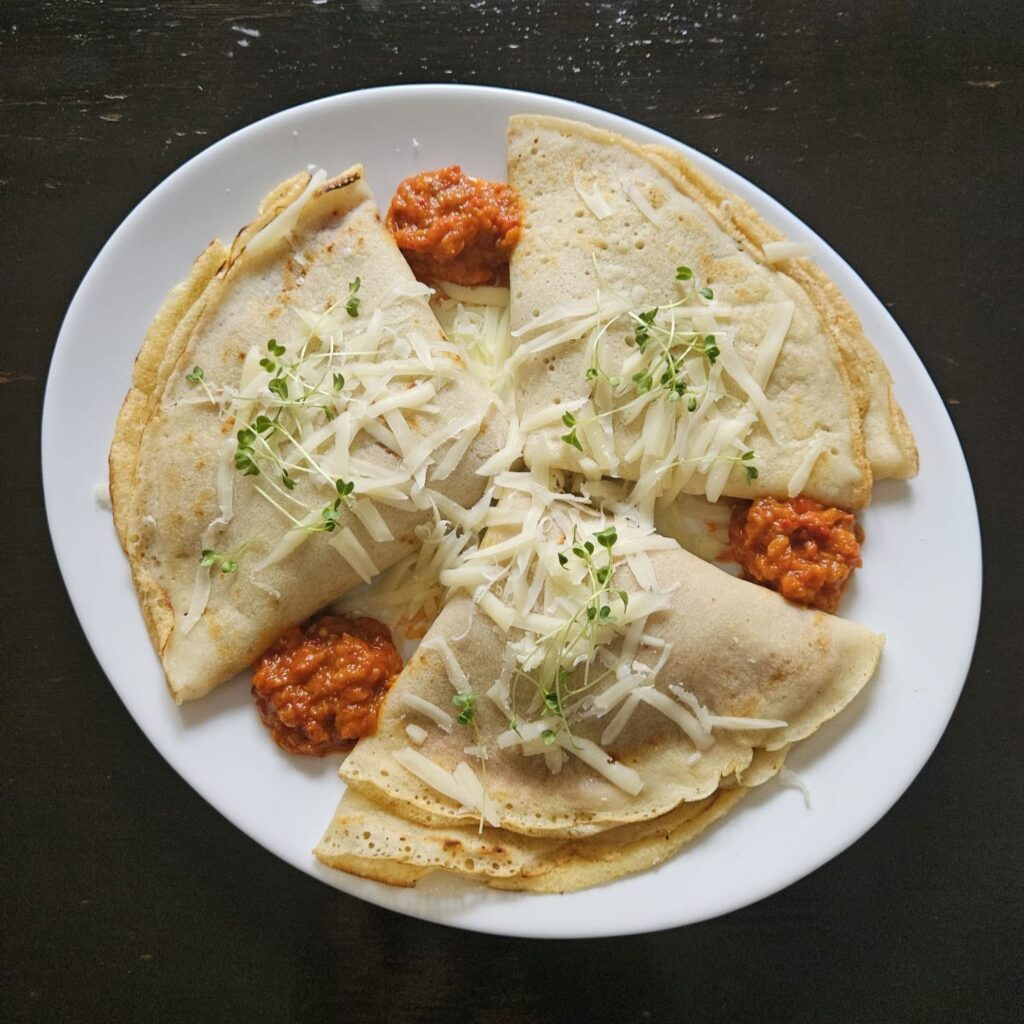 Plate with three ajvar garlic cheese crepes decorated with ajvar, grated cheese, and cabbage microgreens.