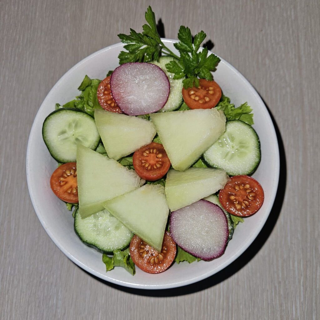 Melon tomato green salad with chopped lettuce, round slices of cucumber and radish, halved cherry tomatoes, and parsley garnish.