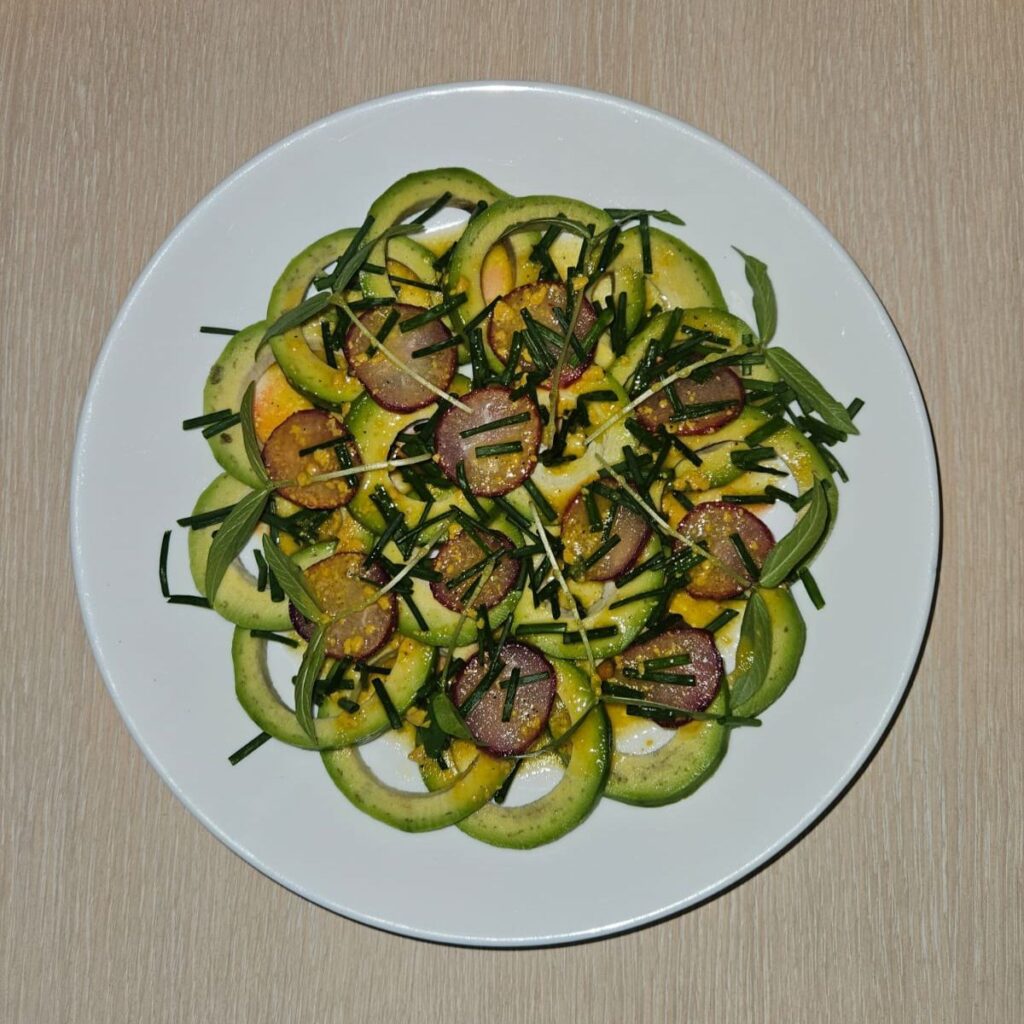 Plated avocado and radish carpaccio drizzled with citrus dressing and topped with mung bean microgreens, served in shallow white bowl.