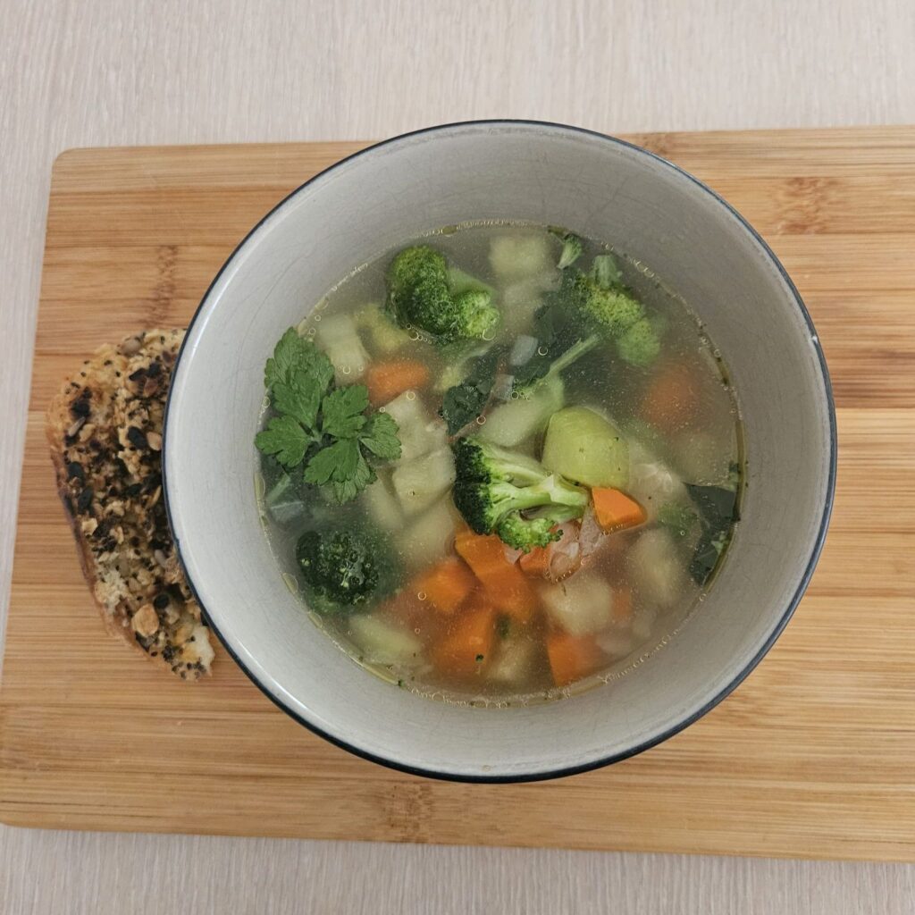 A bowl of warm vegetable soup served on a wooden board beside a slice of whole wheat bread.