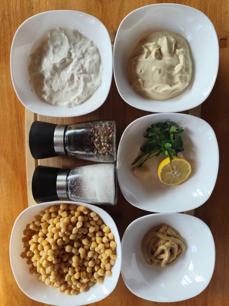 Ingredients for Easy Chickpea Spread Recipe arranged on a table with chickpeas, sour cream, mayonnaise, mustard, lemon, garlic, parsley, salt, and pepper.