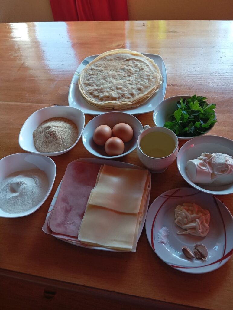 Crepes, ham, cheese, eggs, flour, breadcrumbs, and other ingredients arranged neatly on a table.