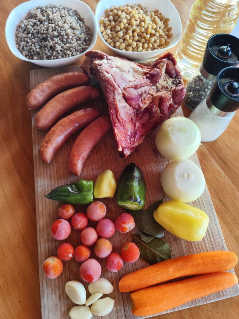 All ingredients for Barley Chickpea Meat Stew laid out on a table, including smoked pork, sausages, chickpeas, barley, vegetables, and spices.