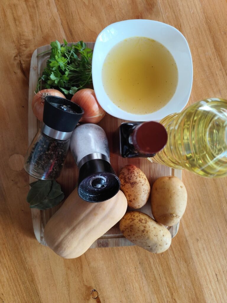All ingredients for Golden Butternut Squash Soup arranged on a table, including butternut squash, onions, potatoes, parsley, bay leaves, oil, and chicken broth.