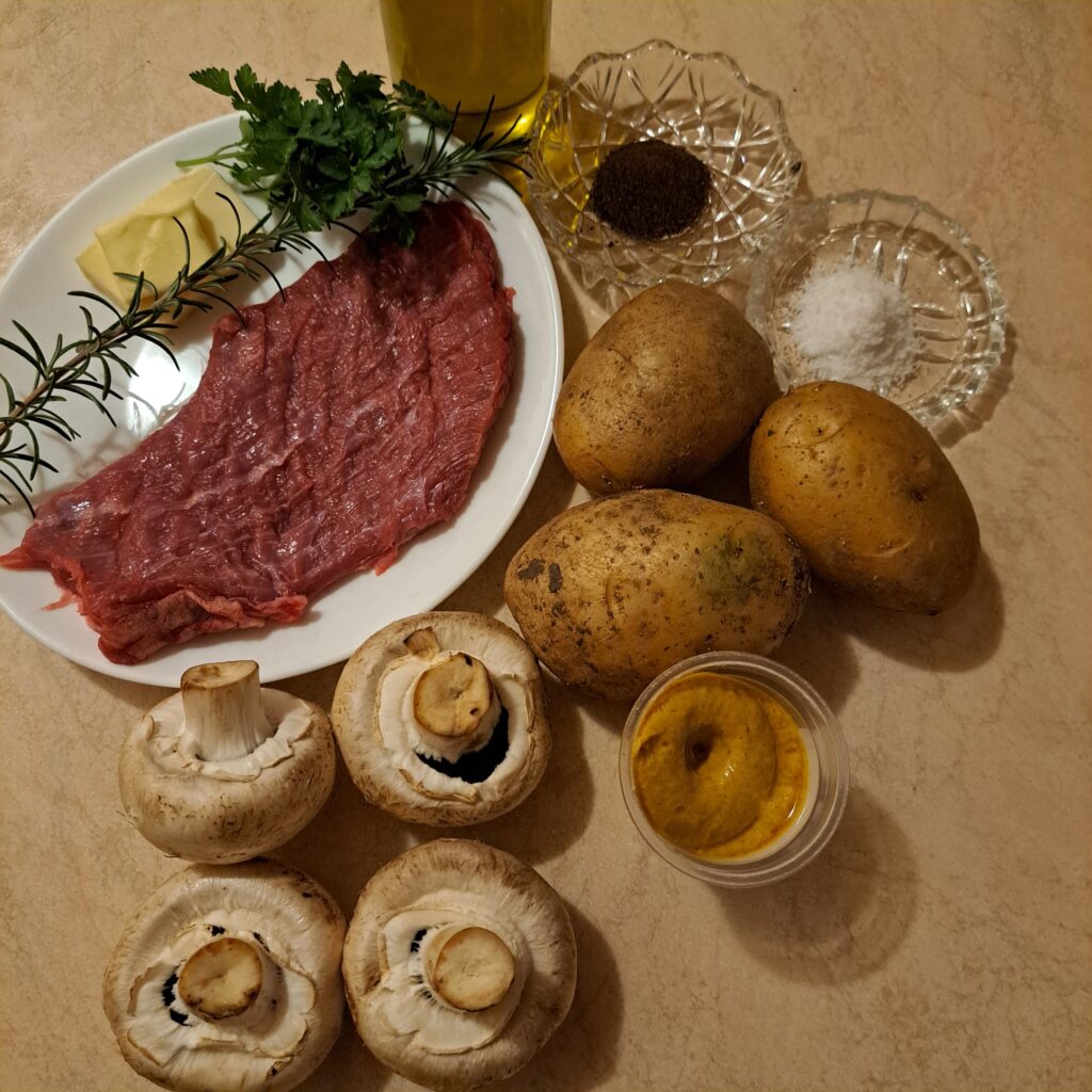 Ingredients for Beef Steak Mushroom Mash on a kitchen table: beef, mushrooms, potatoes, fresh parsley, rosemary, mustard, butter, olive oil, salt, and black pepper.