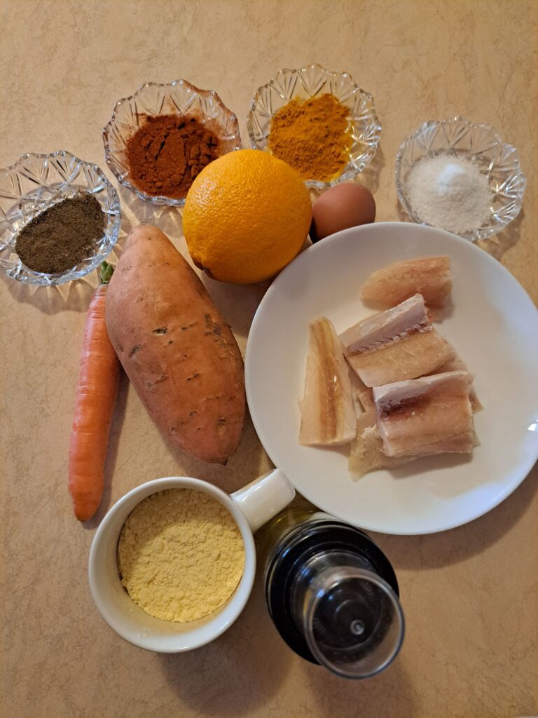 Ingredients on a kitchen table for Sweet Fish Dill Bites including fish fillets, sweet potato, carrot, cornmeal, egg, and spices.