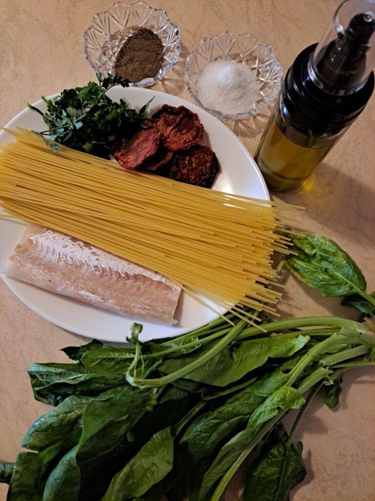 Kitchen table with fresh fish fillet, spaghetti, spinach, sun-dried tomatoes, parsley, olive oil, salt, and black pepper prepared for cooking.