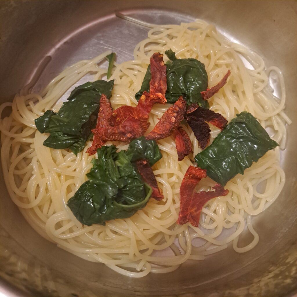 Drained spaghetti in a pot mixed with blanched spinach and sliced sun-dried tomatoes.