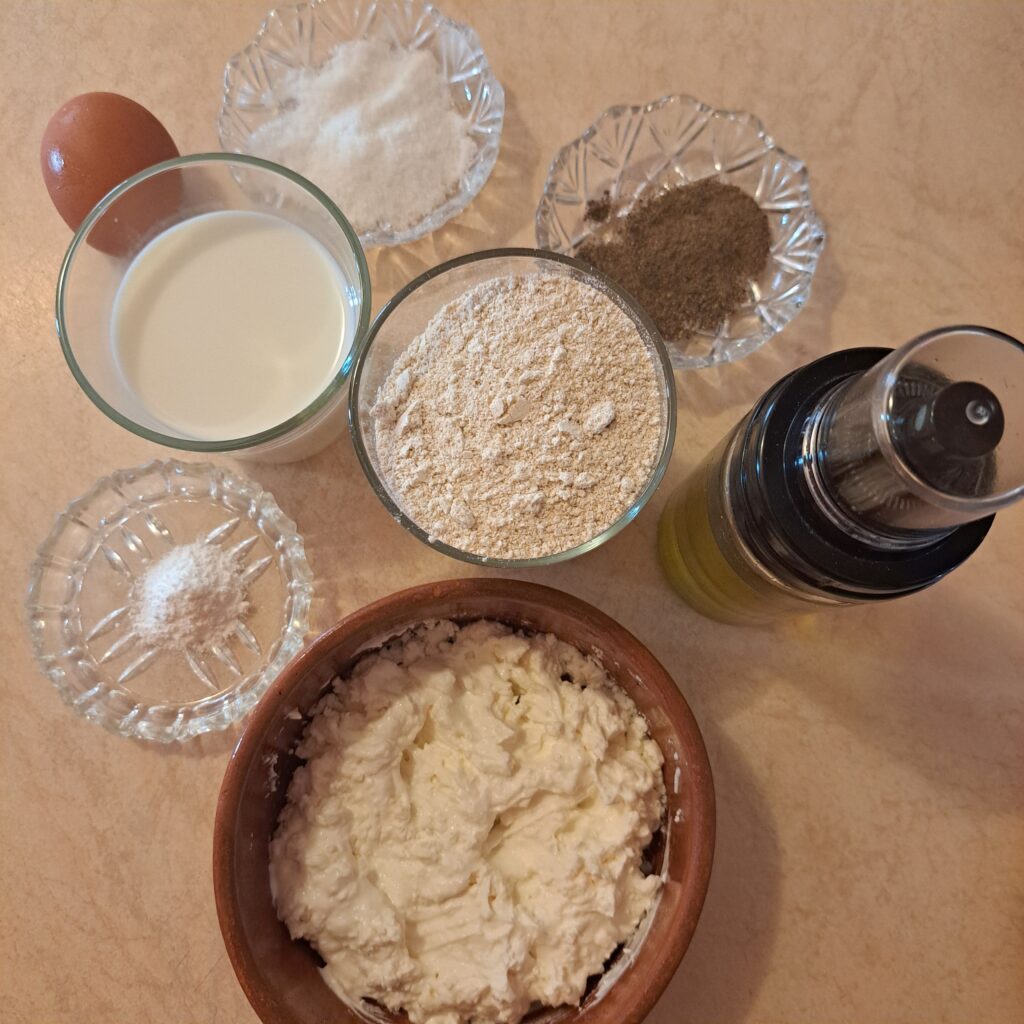 Oat flour, milk, ricotta cheese, egg, salt, black pepper, and olive oil neatly arranged on a kitchen table.
