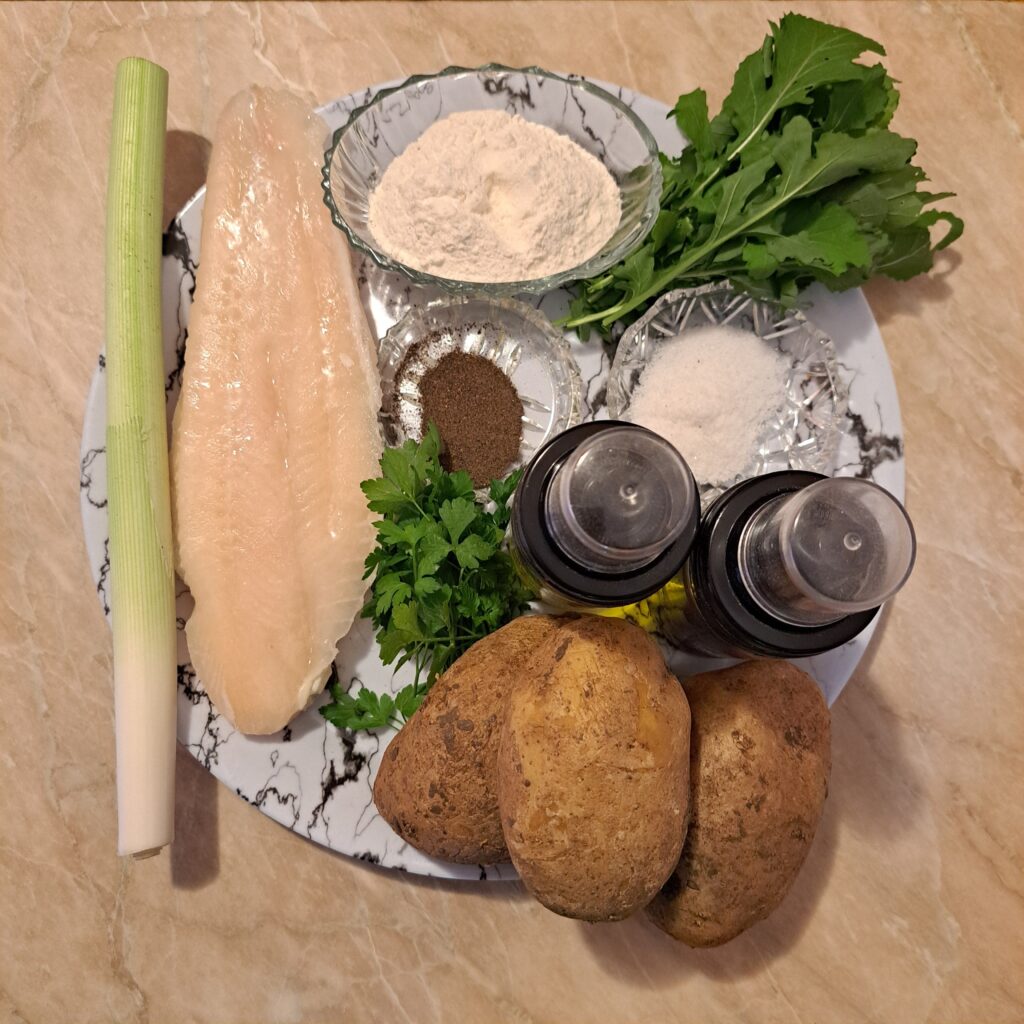 Top view of all ingredients for Fried Pangasius Potato Salad arranged in a wide flat plate, including Pangasius fillets, potatoes, leek, flours, olive oil, apple cider vinegar, salt, and black pepper.