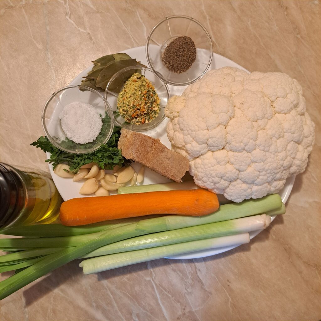 All ingredients for Cauliflower Leek Carrot Soup arranged on a white plate, including cauliflower, leek, carrot, garlic, bread, and seasonings.