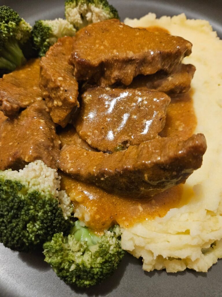 Close-up view of juicy beef slices in brown gravy served with creamy mashed potatoes and lightly blanched broccoli on a plate.