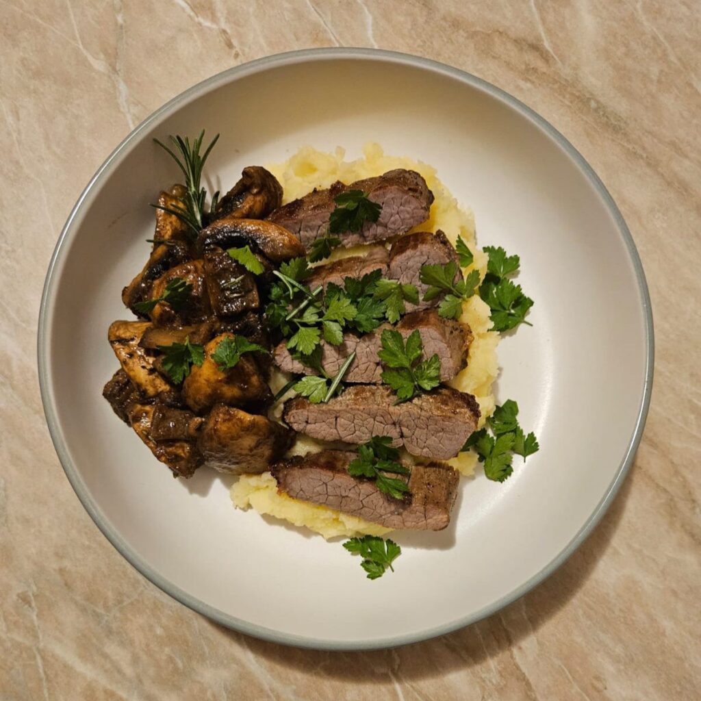Sliced pan-seared beef steak on creamy potato mash, topped with sautéed mushrooms, rosemary, mustard, and chopped fresh parsley.