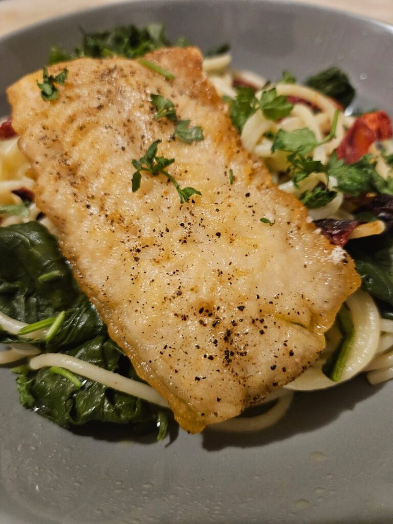 Close-up of the dish Seared Fish Fillet with Pasta and Greens served with spinach, sun-dried tomatoes, and fresh parsley.