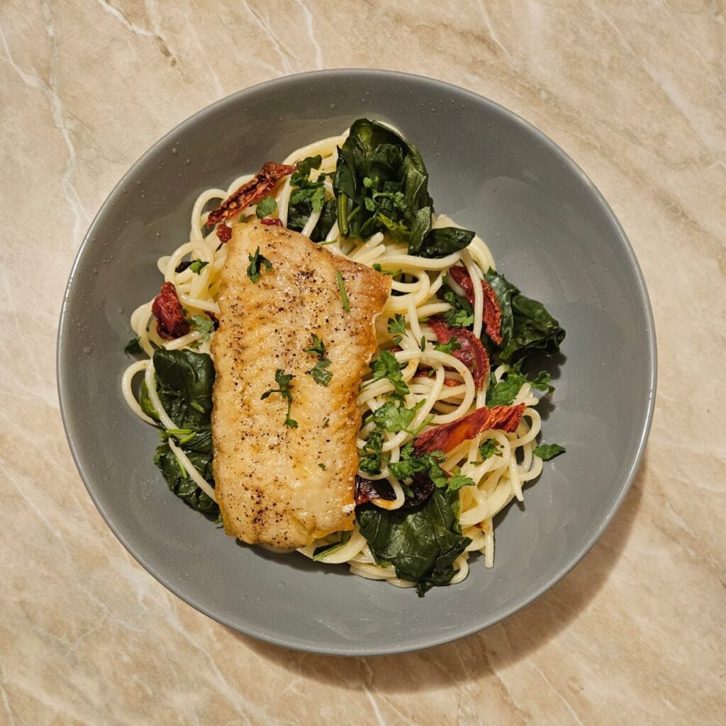 Seared Fish Fillet with Pasta and Greens served on a plate with spinach and sun-dried tomatoes, drizzled with olive oil and sprinkled with chopped parsley.
