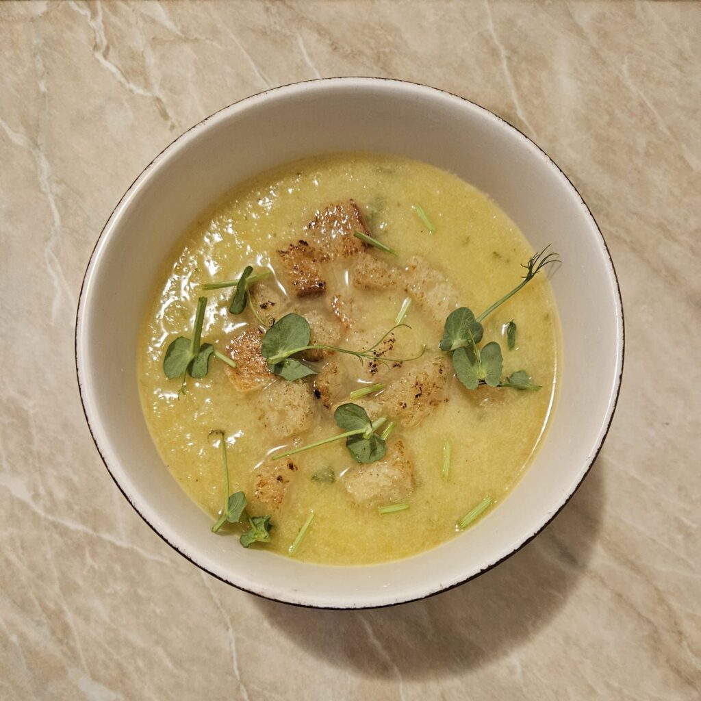 A bowl filled with smooth Cauliflower Leek Carrot Soup, topped with crispy bread cubes and decorated with fresh pea microgreens.