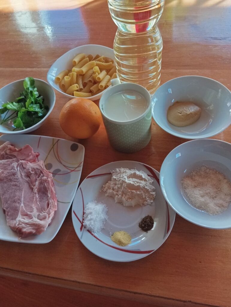 Ingredients for Creamy Pork Neck Rigatoni arranged on a kitchen table, including pork neck, rigatoni, milk, orange, mustard, flour, and parsley.
