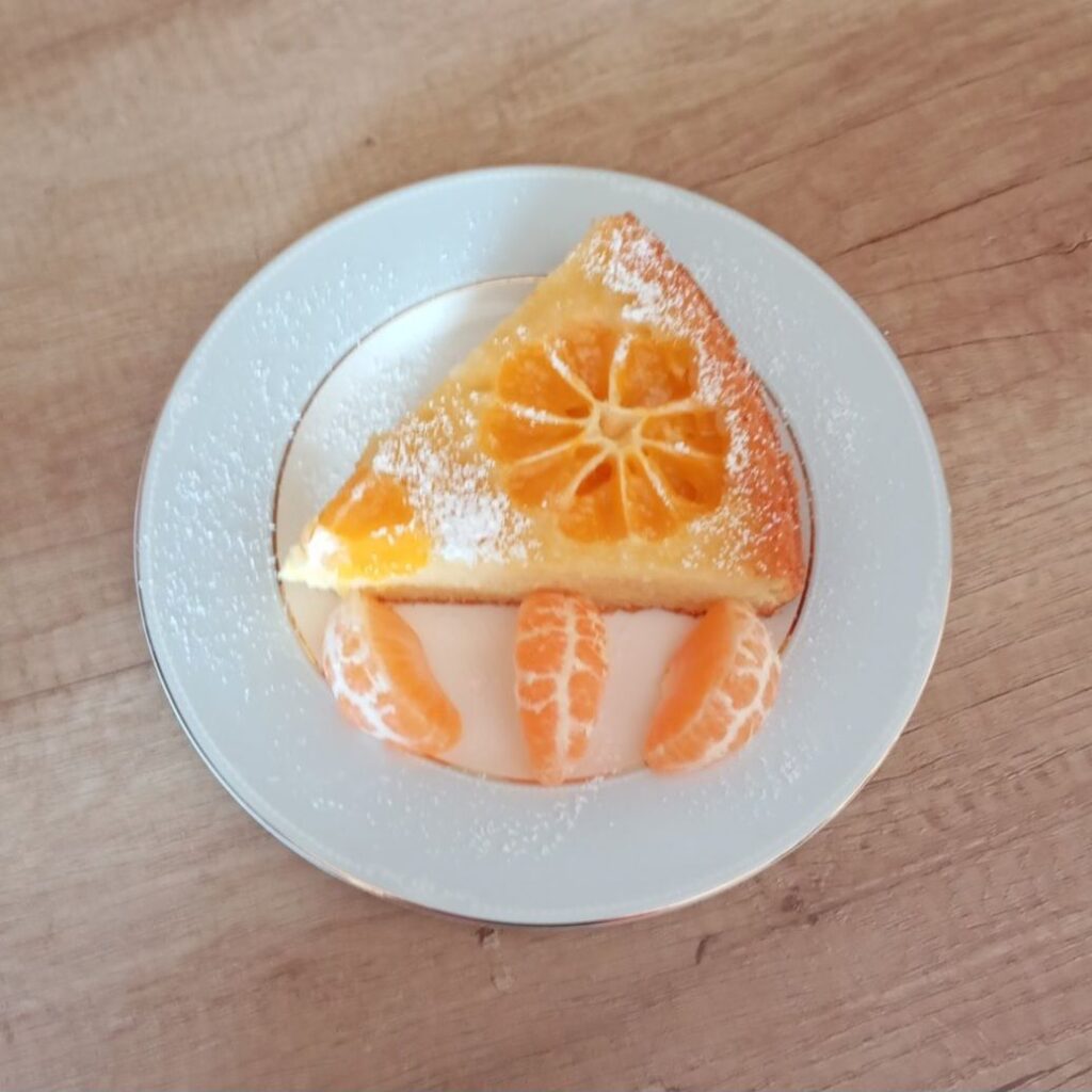 A single slice of Simple Juicy Mandarin Cake on a plate, dusted with powdered sugar and accompanied by a fresh mandarin slice.