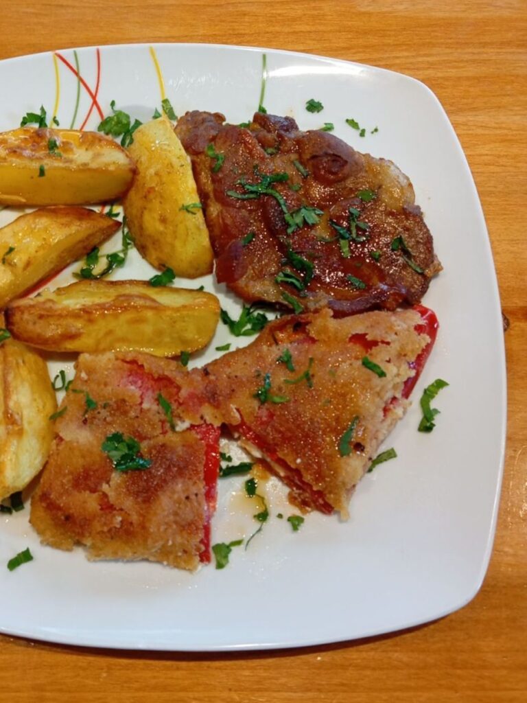 Close-up of a plate with one boneless pork neck medallion, a halved stuffed red pepper, and golden roasted potatoes.