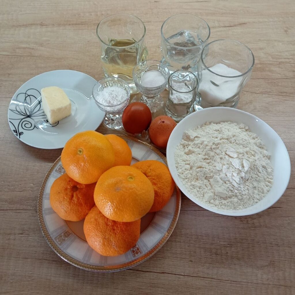 Fresh ingredients for Simple Juicy Mandarin Cake arranged on a kitchen table, including eggs, mandarins, flour, sugar, oil, and butter.