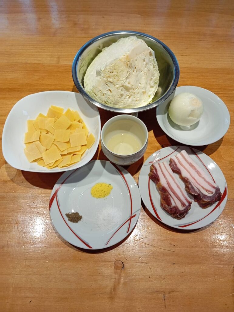 Top view of ingredients for Bacon Cabbage Pasta Squares arranged on a kitchen table, including cabbage, onion, oil, egg pasta squares, bacon, and seasonings.
