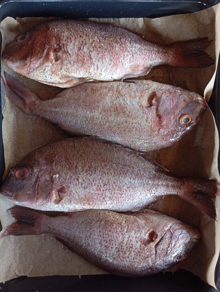 Four whole red seabream arranged on baking paper in a tray, seasoned with salt, fish seasoning, and black pepper, ready for Baked Red Seabream Delight.