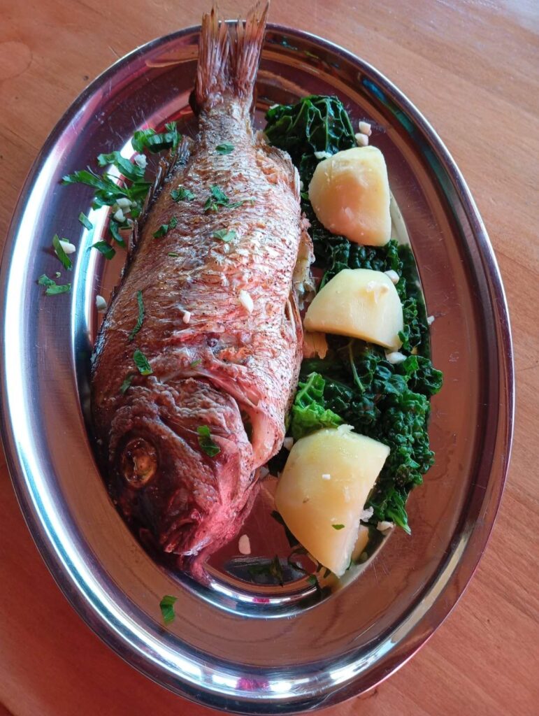Baked red seabream served in an oval dish with boiled potato pieces and cooked kale.