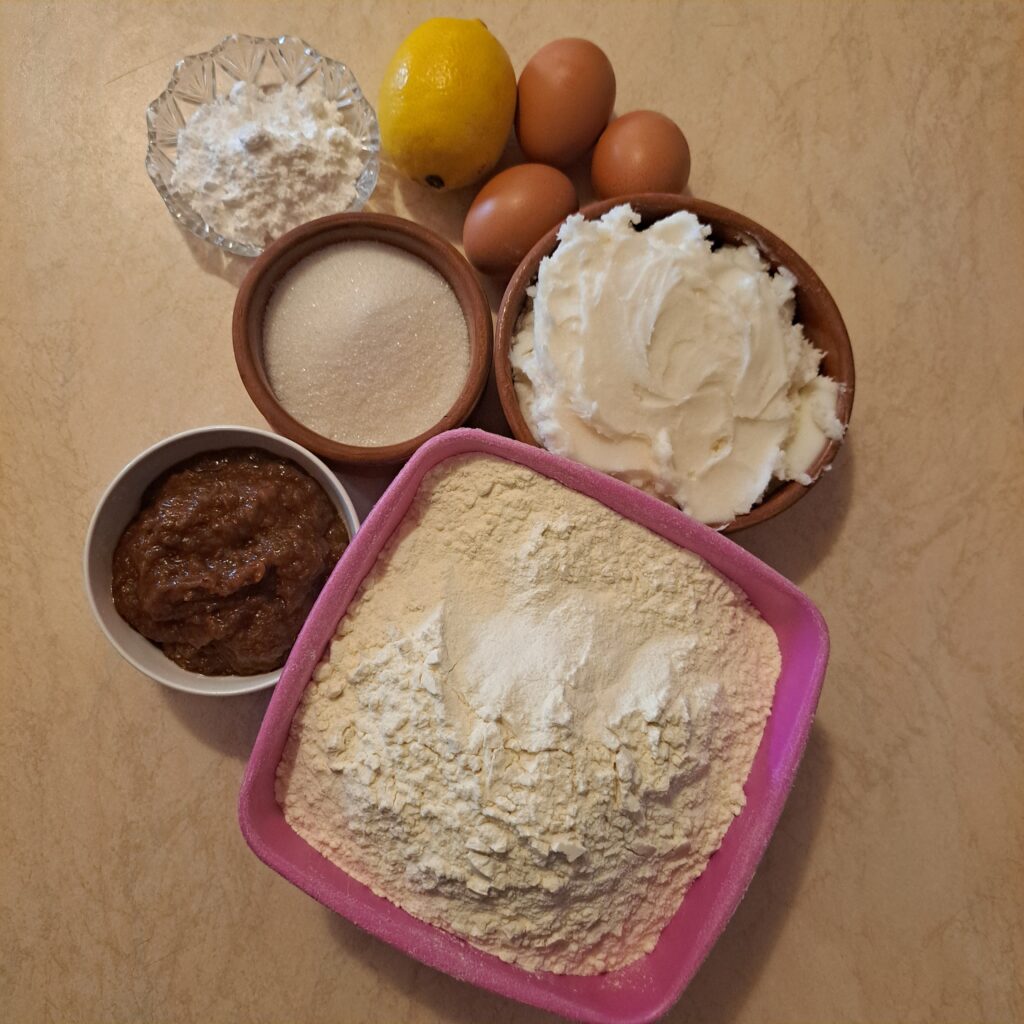 All ingredients for Baked Jam Sandwich Cookies arranged on a kitchen table, including flour, lard, sugar, eggs, lemon, powdered sugar, vanilla, and plum jam.