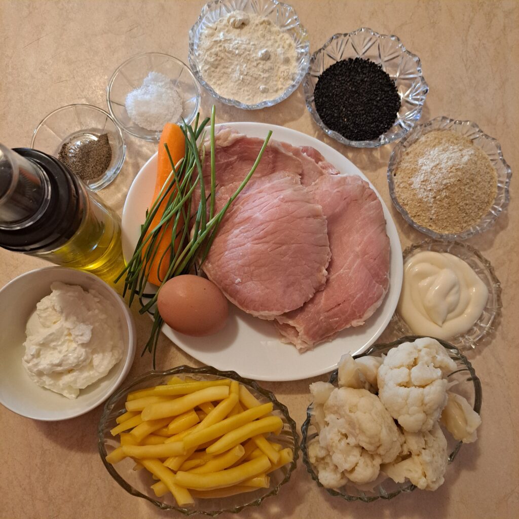 Ingredients for Crispy Pork Veggie Plate arranged on a kitchen counter, including pork schnitzels, green beans, cauliflower, eggs, breadcrumbs, and sauce components.