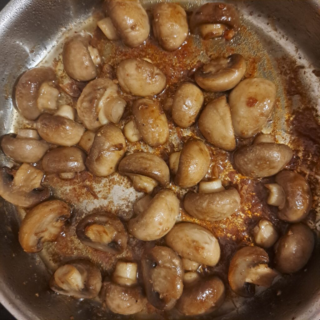 Halved mushrooms frying in the same pan used for chicken, with garlic added and lightly seasoned.