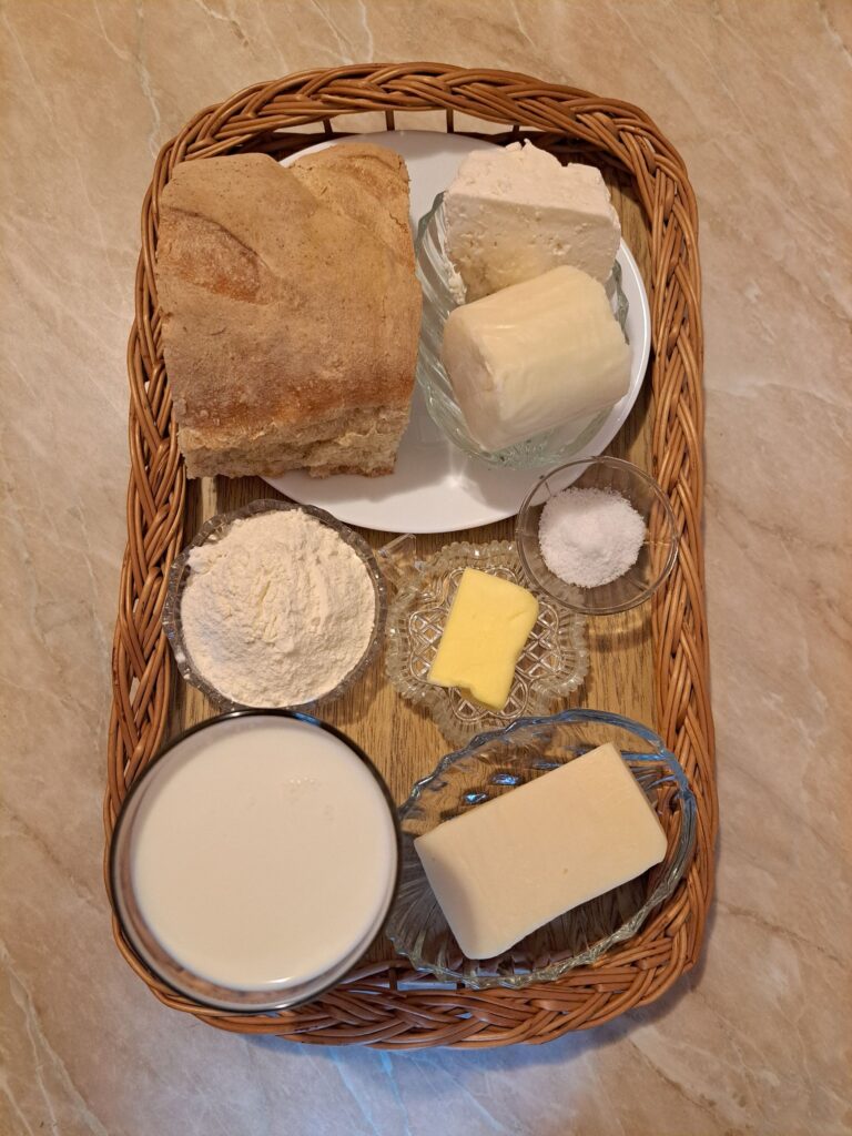 Stale bread, butter, flour, milk, salt, and whole pieces of hard cheese, yellow cheese, and mozzarella arranged neatly in a flat wooden tray.