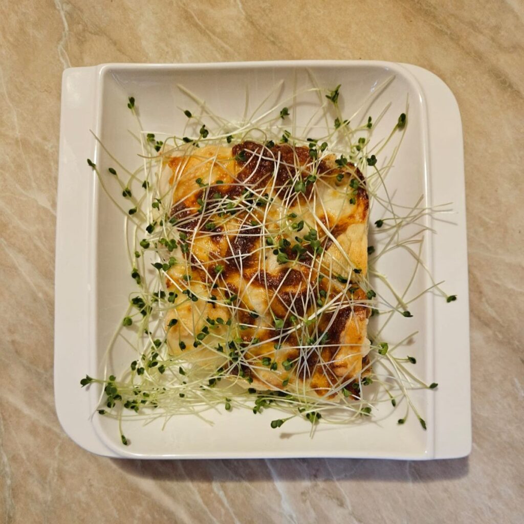 A plated portion of Baked Cheesy Bread Cubes topped generously with fresh broccoli microgreens, showing golden melted cheese and creamy béchamel layers.