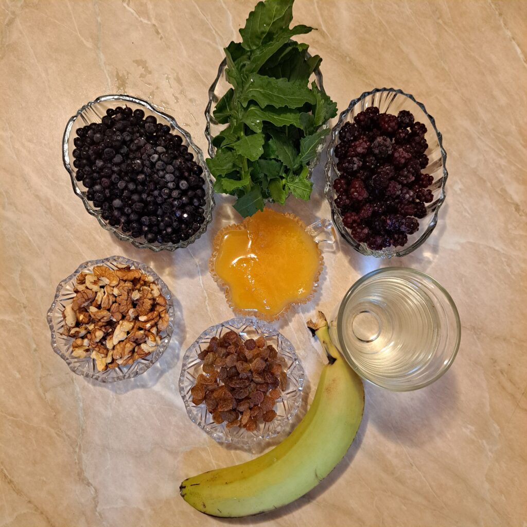 All ingredients arranged on a table: frozen blackberries and blueberries, honey, banana, raisins, arugula, and walnuts.